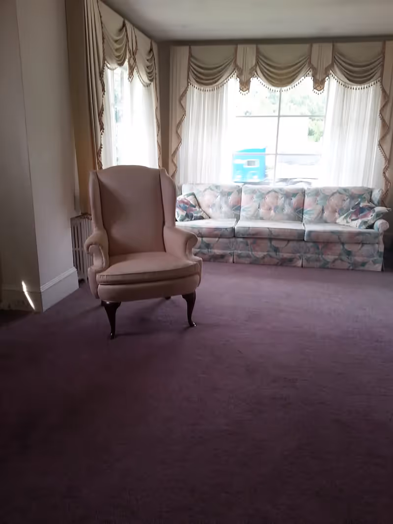 A living room with a large window covered by sheer curtains and decorative valances. In front of the window is a floral-patterned sofa with two matching cushions. A single upholstered armchair with wooden legs is positioned on a purple carpeted floor.