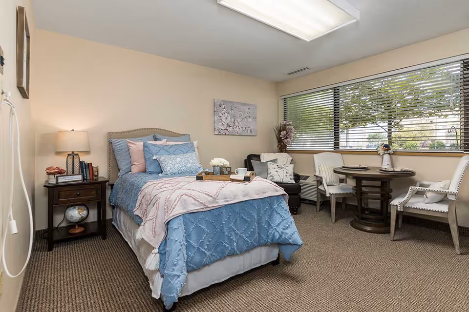 Furnished bedroom with a made bed, nightstand, lamp, and a seating/dining area by a large window with blinds.