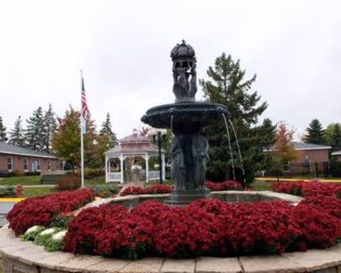 A tiered fountain surrounded by red flowers in a landscaped courtyard with a white gazebo and an American flag.