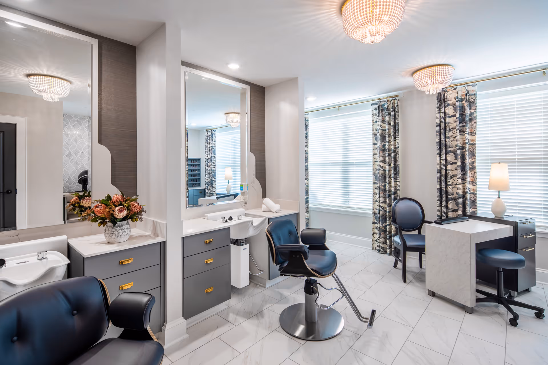 A bright and modern salon area with two black salon chairs in front of large mirrors and gray cabinetry. There is a small white sink on the left side, a vase with flowers on the counter, and a small desk with two chairs near large windows with patterned curtains. The floor is white tile, and two elegant ceiling lights illuminate the room.