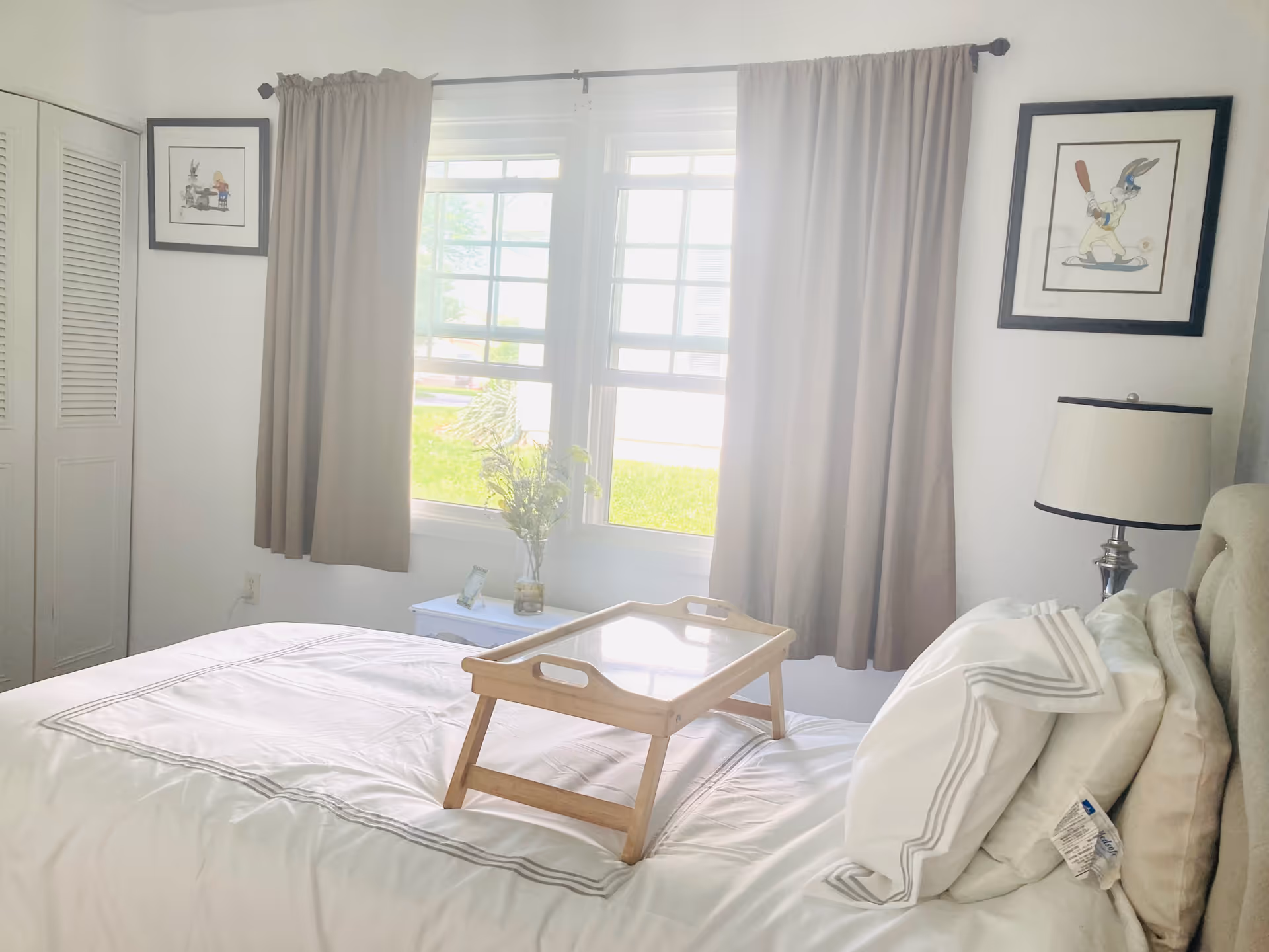 A bright bedroom with a neatly made bed featuring white linens and pillows. A wooden tray is placed on the bed. There is a window with beige curtains letting in natural light. On either side of the window are framed pictures, and a bedside table with a lamp and a small vase with flowers is visible.