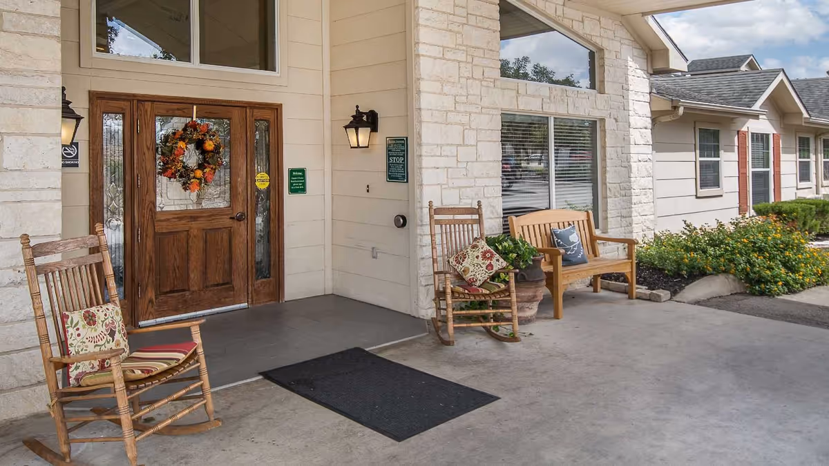 Entrance to a building with wooden double doors decorated with a fall-themed wreath. Two wooden rocking chairs with patterned cushions and a wooden bench with a cushion are placed on the concrete porch. There are stone walls, a window with blinds, and some greenery and flowers near the building.