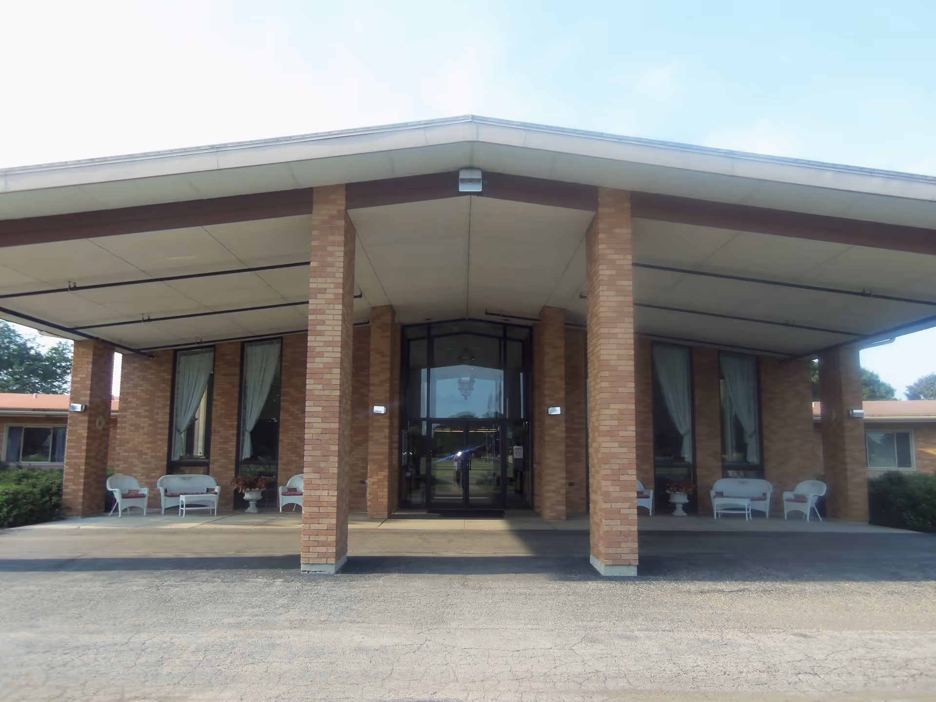 Front entrance of a brick senior living facility with a covered drop-off, columns, glass doors, and outdoor seating.