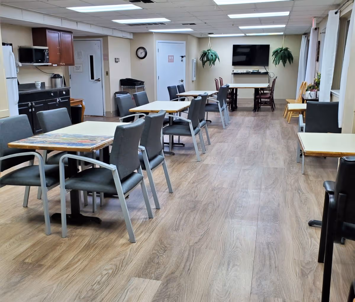 Interior view of a senior living facility common dining area with multiple tables and chairs arranged neatly. The room has wood flooring, a wall-mounted TV, hanging plants, and a kitchenette area with a microwave, refrigerator, and cabinets. There is a door with restroom signs in the background.