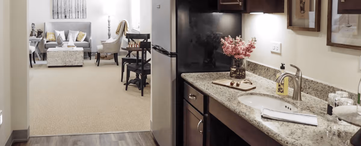 View from a kitchen area with granite countertops and a sink looking into a living room furnished with a sofa, armchair, coffee table, and side tables. The kitchen has dark wood cabinets and a black refrigerator. The living room features neutral tones with decorative pillows and a floor lamp.