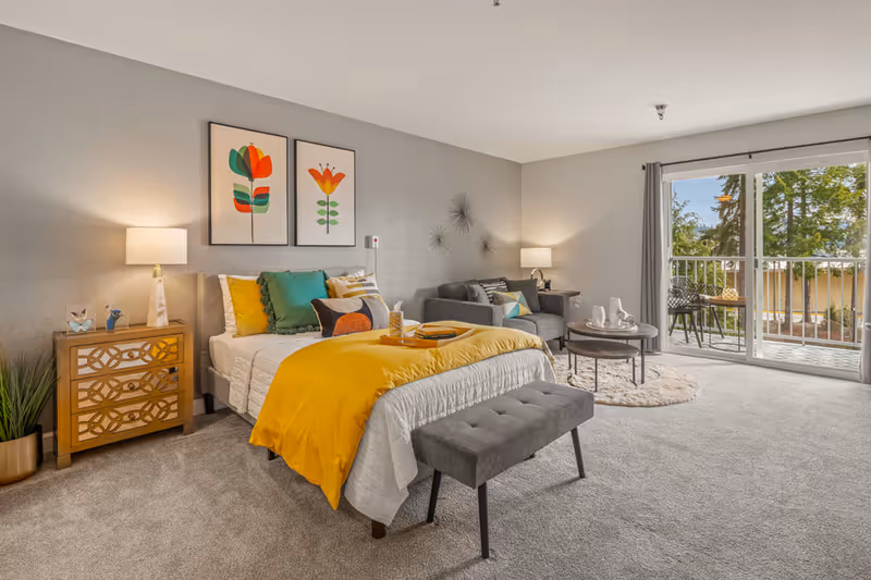 A bright and spacious bedroom with a bed featuring white and yellow bedding, colorful pillows, and a gray bench at the foot. There is a wooden nightstand with a lamp and decorative items on the left side of the bed. Two framed floral artworks hang above the bed. To the right, there is a gray sofa with cushions, a round coffee table on a circular rug, and a sliding glass door leading to a balcony with outdoor chairs and a table. The room has light gray walls and carpeted flooring.