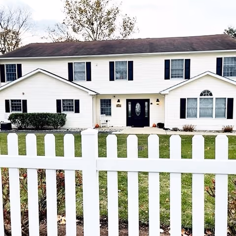 A two-story white residential building with black shutters and a dark roof, viewed from behind a white picket fence. The building has multiple windows and a central entrance door with glass panels. There is a small lawn and some shrubs in front of the building under a cloudy sky.