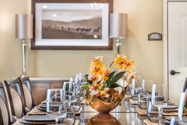 A formal dining table set with glassware and a gold vase of yellow orchids as a centerpiece in a well-appointed dining room.