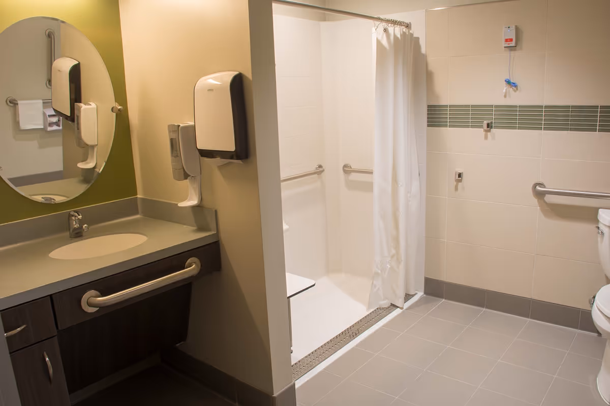 Accessible bathroom with a sink and countertop, a round mirror above the sink, soap and paper towel dispensers mounted on the wall, a walk-in shower with grab bars and a white shower curtain, and a toilet with a grab bar beside it.