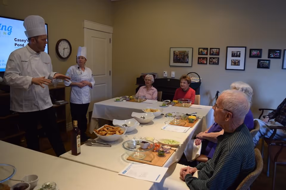 Two chefs in white uniforms and tall hats are conducting a cooking demonstration for a group of elderly people seated around a table with various food ingredients and bowls. The setting is an indoor room with framed photos on the wall and a clock above a door.