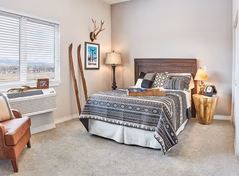 A cozy bedroom with a bed covered in a patterned blue and white blanket, two pillows, and a wooden headboard. There is a wooden side table with a lamp and a clock, a floor lamp next to the bed, and a pair of vintage wooden skis leaning against the wall. A window with blinds lets in natural light, and a brown leather chair is positioned near the window. The room has beige walls and carpeted flooring.