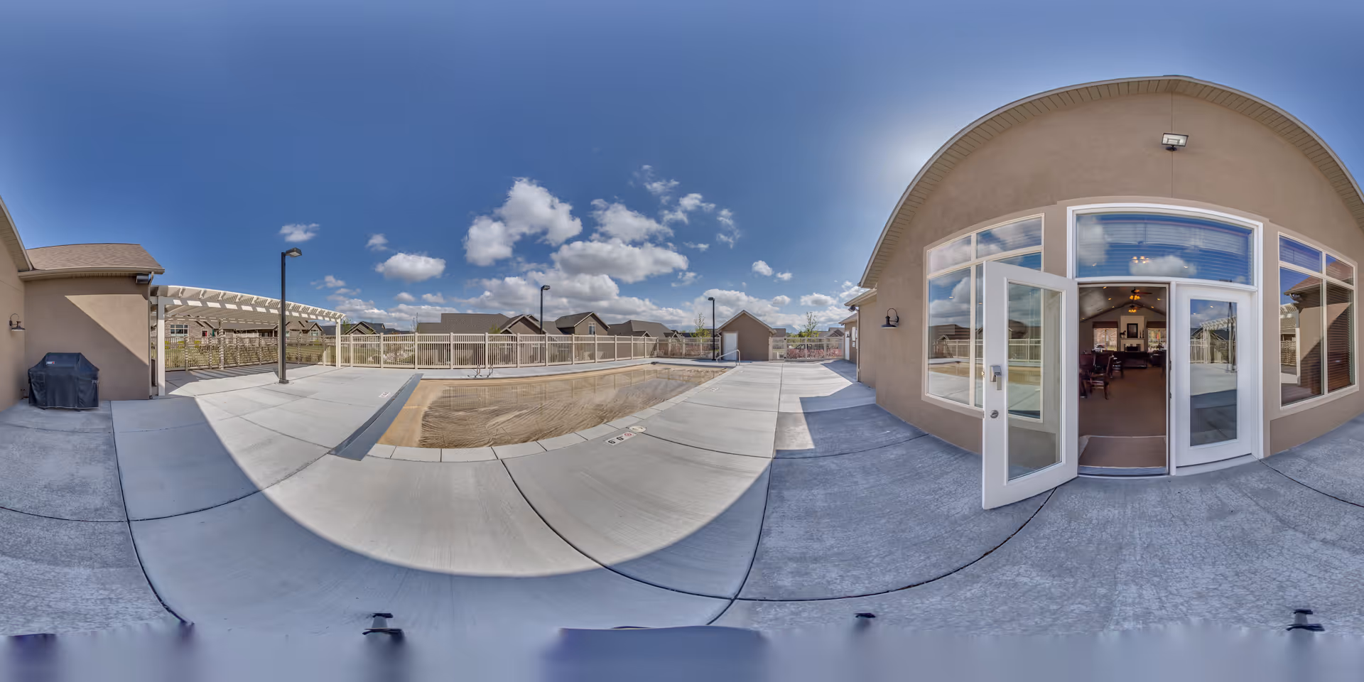 Outdoor area of Bridlewood Villas featuring a fenced empty swimming pool surrounded by concrete pavement, adjacent to a building with large windows and an open door leading inside. The sky is clear with scattered clouds.
