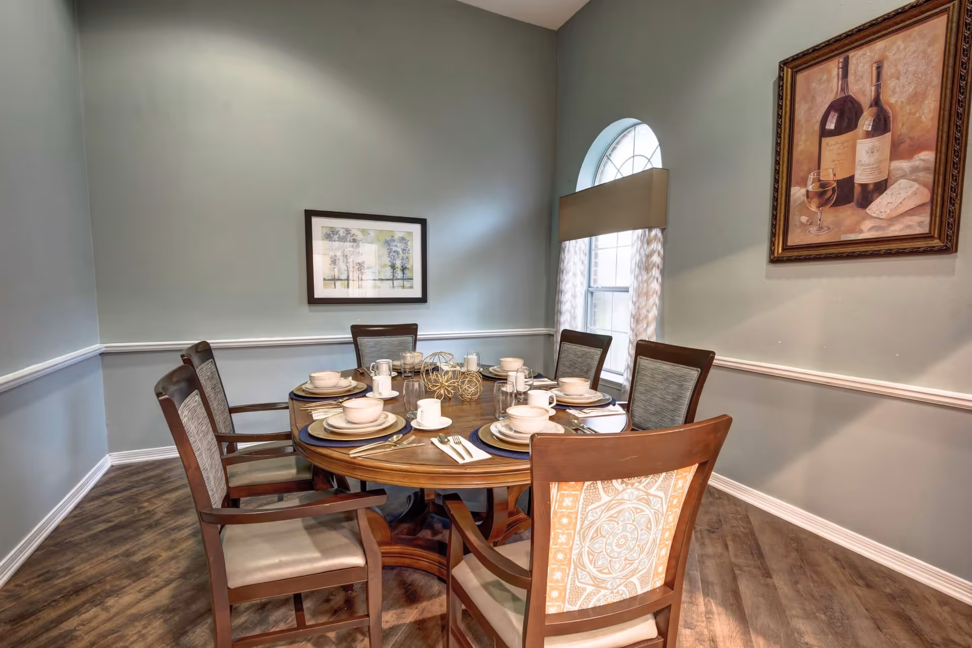A dining room with a round wooden table set for six people with plates, bowls, cups, and silverware. The room has light green walls with white trim, a window with an arched top and patterned curtains, and two framed pictures on the walls, one depicting wine bottles and glasses and the other a landscape.