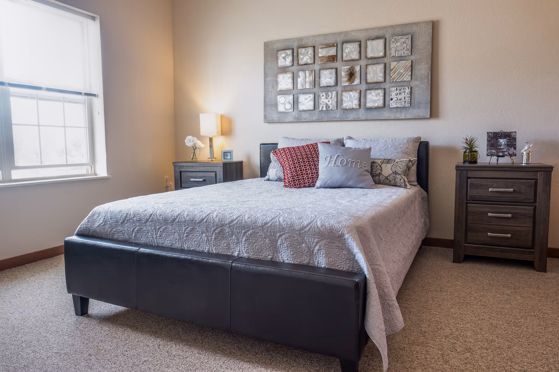 A neatly made bed with decorative pillows between two nightstands beneath wall art in a bright bedroom with a window on the left.