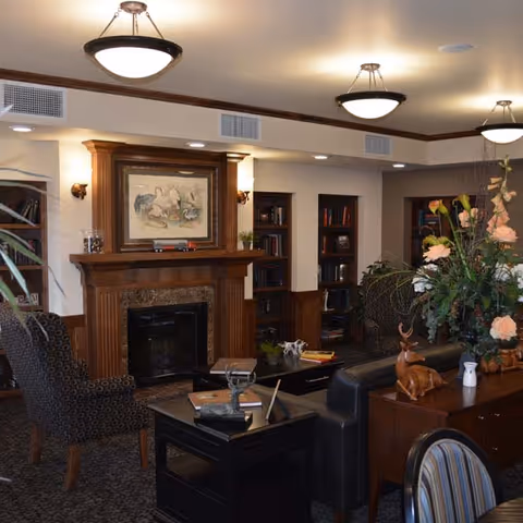 A cozy living room area with a wooden fireplace mantel, framed artwork above the fireplace, built-in bookshelves filled with books, patterned armchairs, a black leather sofa, and a wooden table adorned with decorative items and a large floral arrangement.