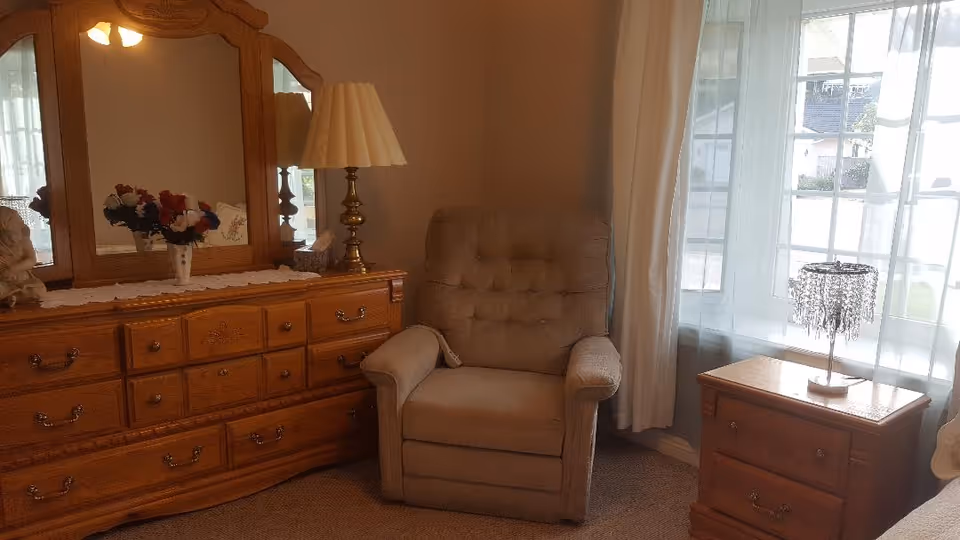A cozy corner of a bedroom featuring a beige upholstered armchair next to a wooden dresser with a large mirror, a table lamp, and a vase of flowers. To the right, there is a wooden nightstand with a decorative lamp in front of a window with sheer white curtains.