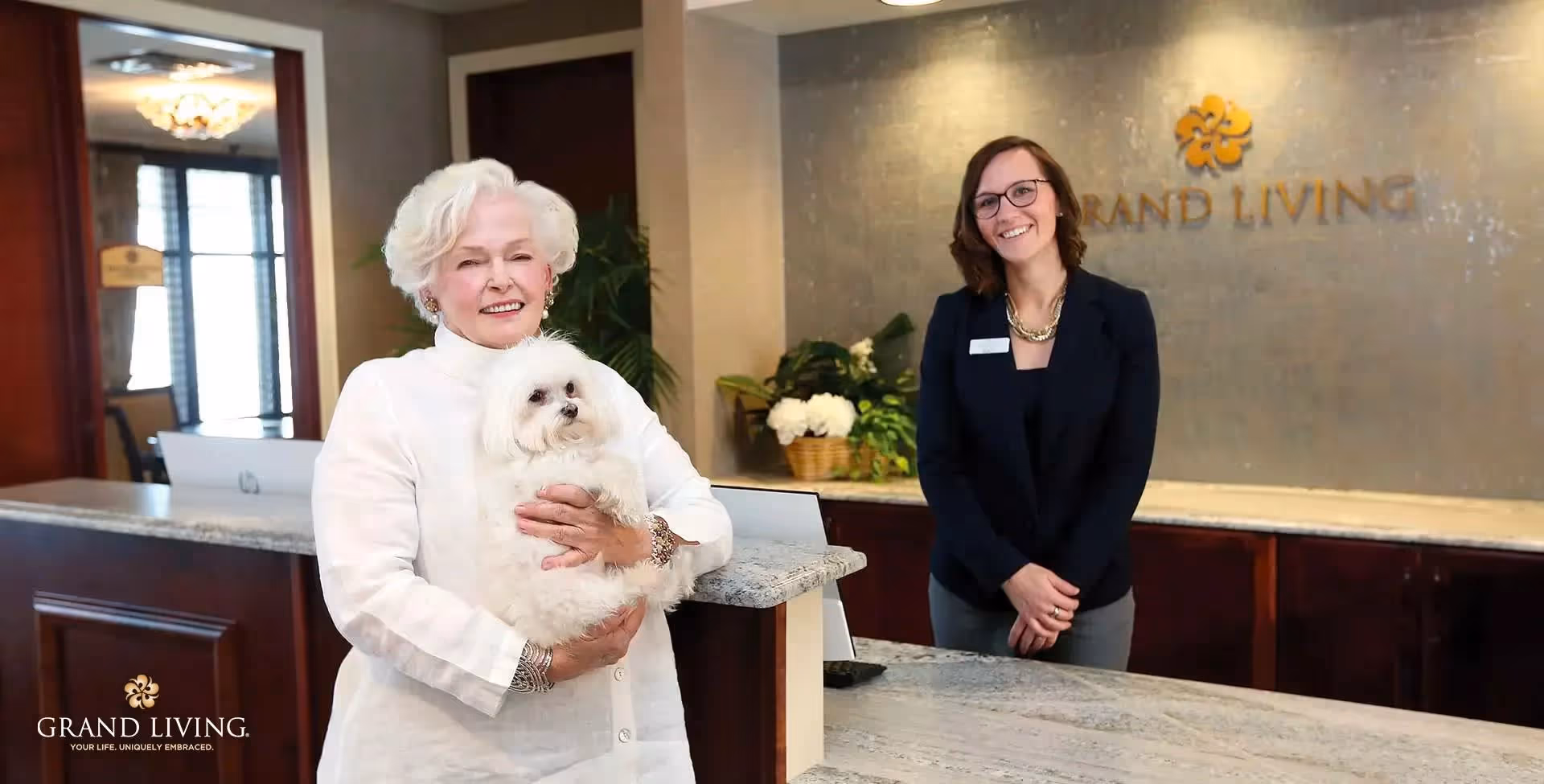 An elderly woman holding a small white dog stands smiling in front of a reception desk. Behind the desk, a smiling woman wearing glasses and a dark blazer stands with her hands clasped. The background features a wall with the Grand Living logo and some plants.