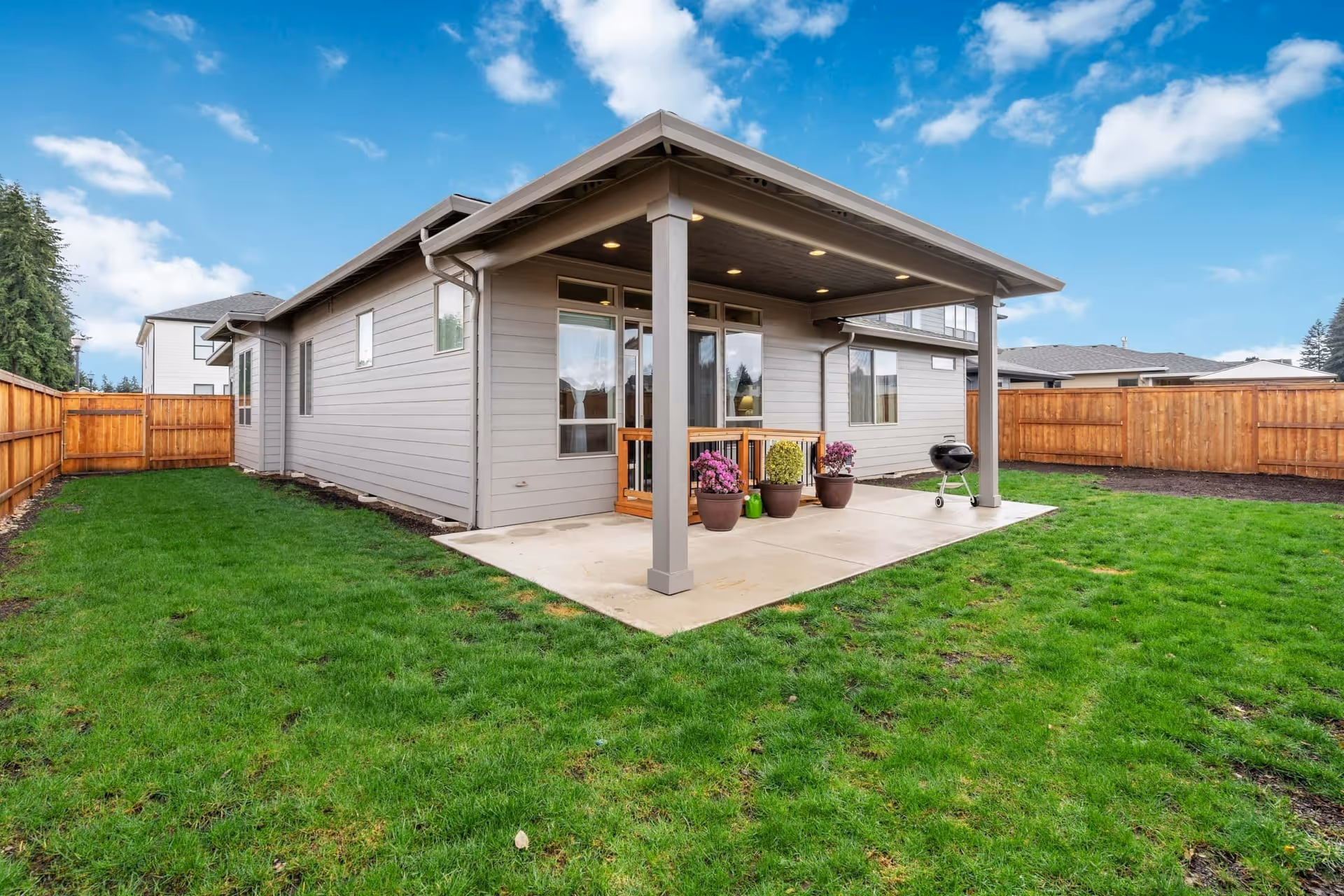 Backyard view of a single-story gray house with a covered patio, potted plants, a small grill, and a fenced green lawn under a blue sky.