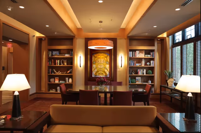 A cozy and well-lit living room area with a beige sofa in the foreground, two table lamps on side tables, built-in wooden bookshelves filled with books and decorative items, a glass-top table with chairs in the center, a modern circular ceiling light fixture, and large windows with wooden blinds on the right side.