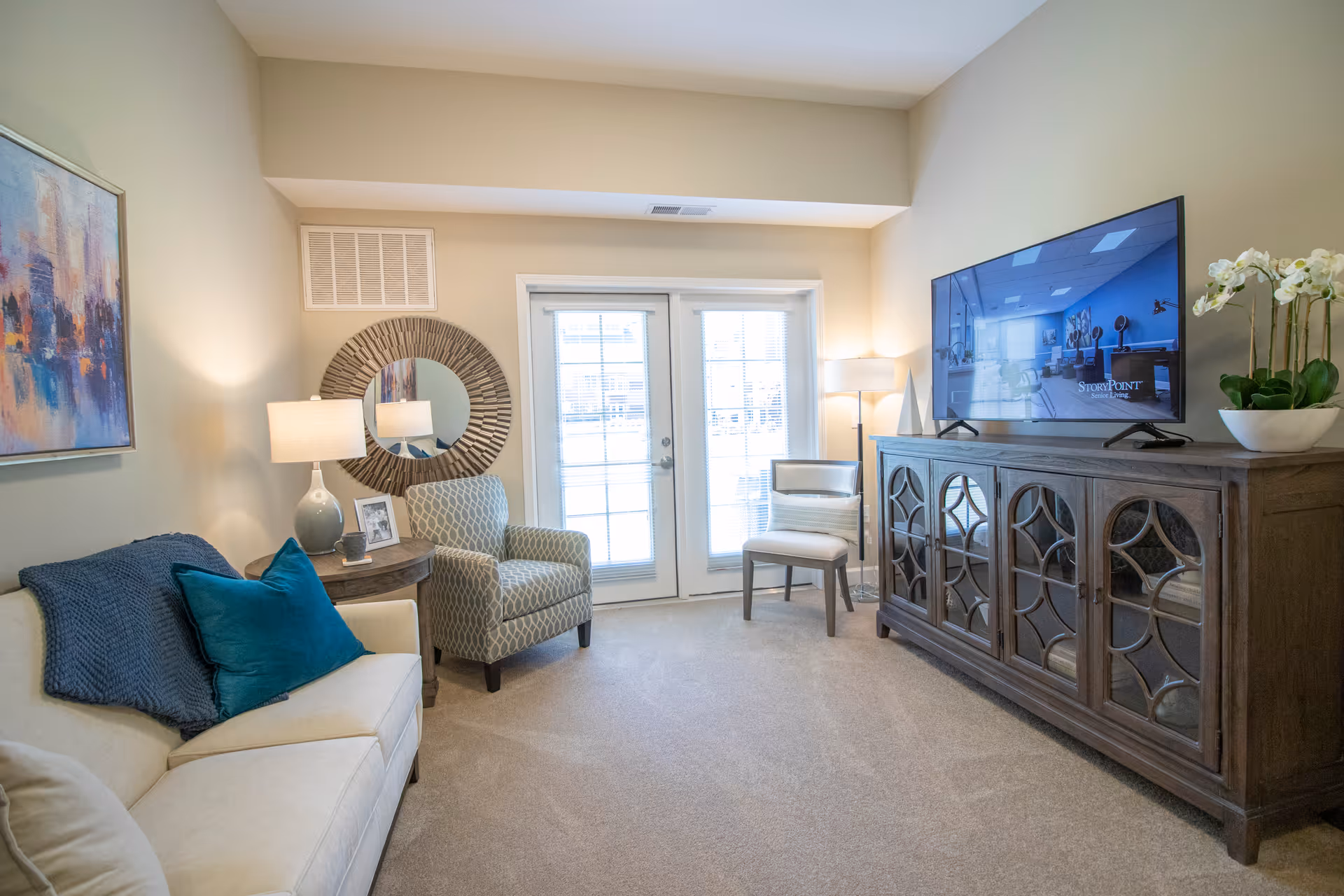 A cozy living room with a beige sofa adorned with a blue throw blanket and teal pillow, a patterned armchair, a wooden side table with a lamp and framed photo, a round decorative mirror on the wall, a wooden cabinet with glass doors holding a flat-screen TV displaying the StoryPoint logo, a white chair, a floor lamp, and a potted orchid plant. French doors with blinds allow natural light into the room.