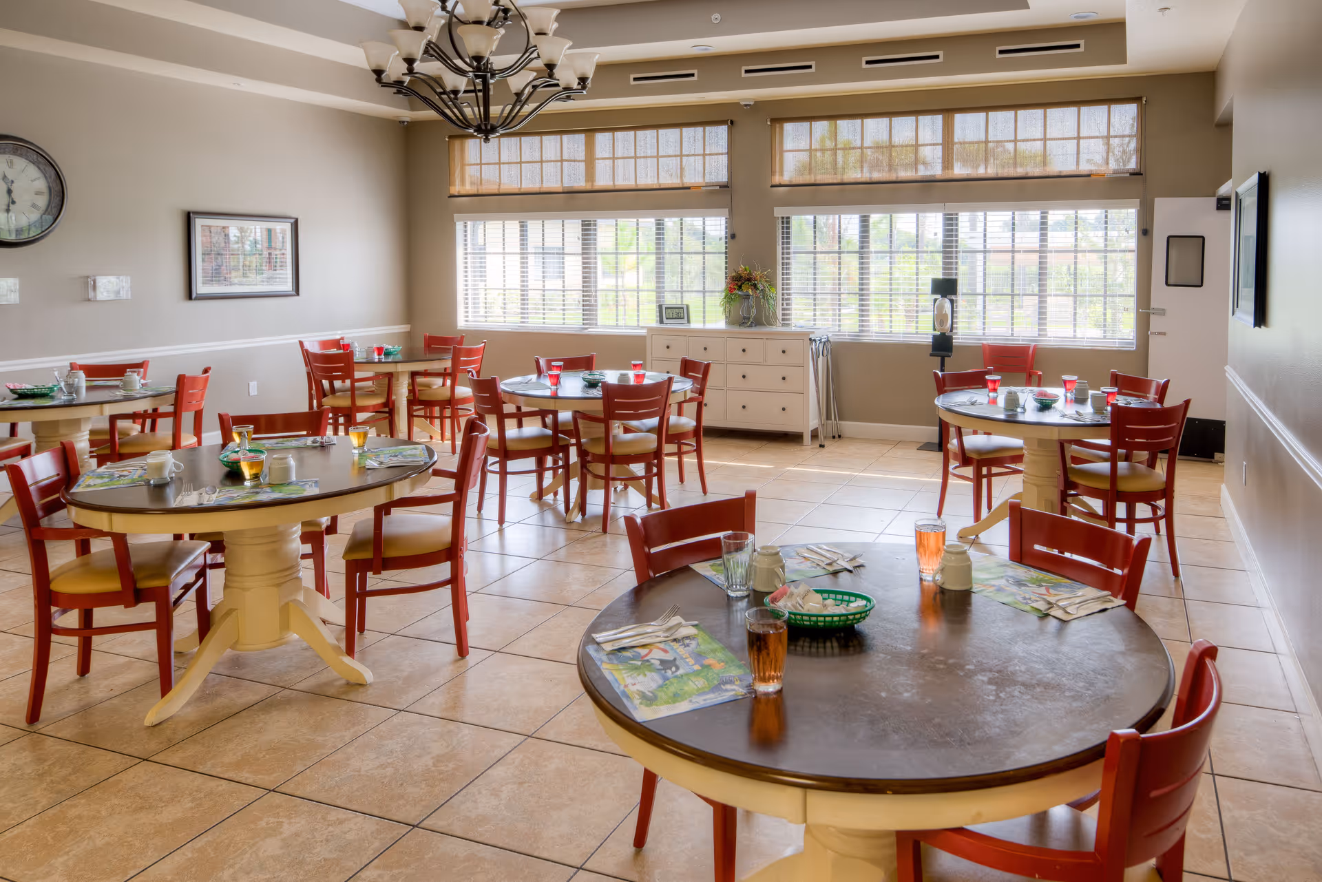 A bright dining room with multiple round tables set with placemats, glasses, and utensils. The room has large windows with blinds allowing natural light to fill the space. Red chairs surround the tables, and a chandelier hangs from the ceiling. The walls are painted beige with a clock and framed picture on one side.