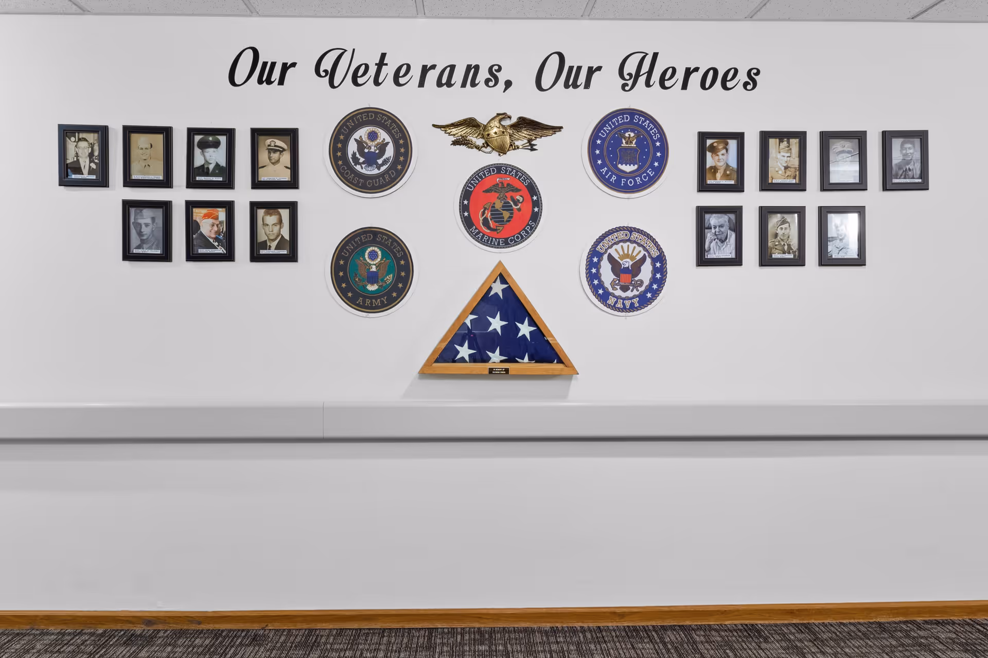 A hallway wall display honoring veterans with framed photos, military service emblems, and a folded American flag beneath the heading "Our Veterans, Our Heroes".