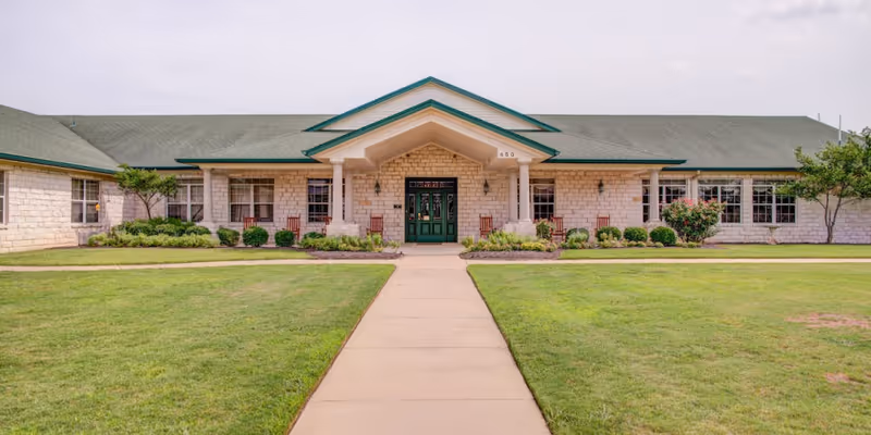 Single-story stone building front with a covered entrance, green double doors, and a walkway across a manicured lawn.