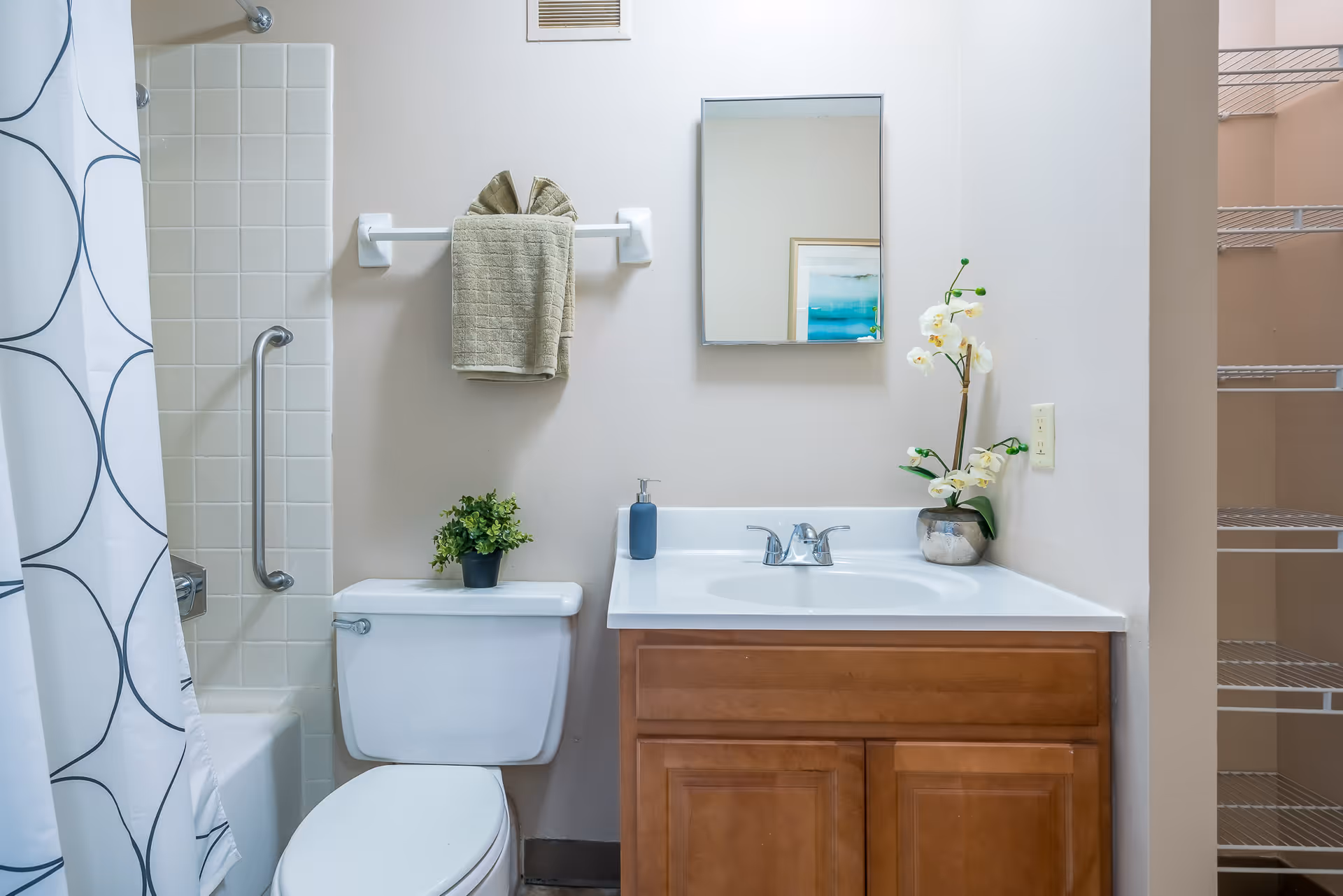 Bathroom with a toilet next to a wooden vanity and sink, mirror above, a shower with patterned curtain to the left, and towels and plants as decor.