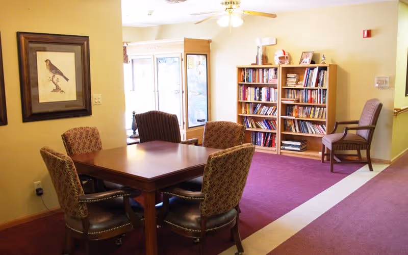 A cozy common area with a wooden table surrounded by four patterned chairs, a bookshelf filled with books against a yellow wall, a framed bird picture on the wall, and a window letting in natural light.