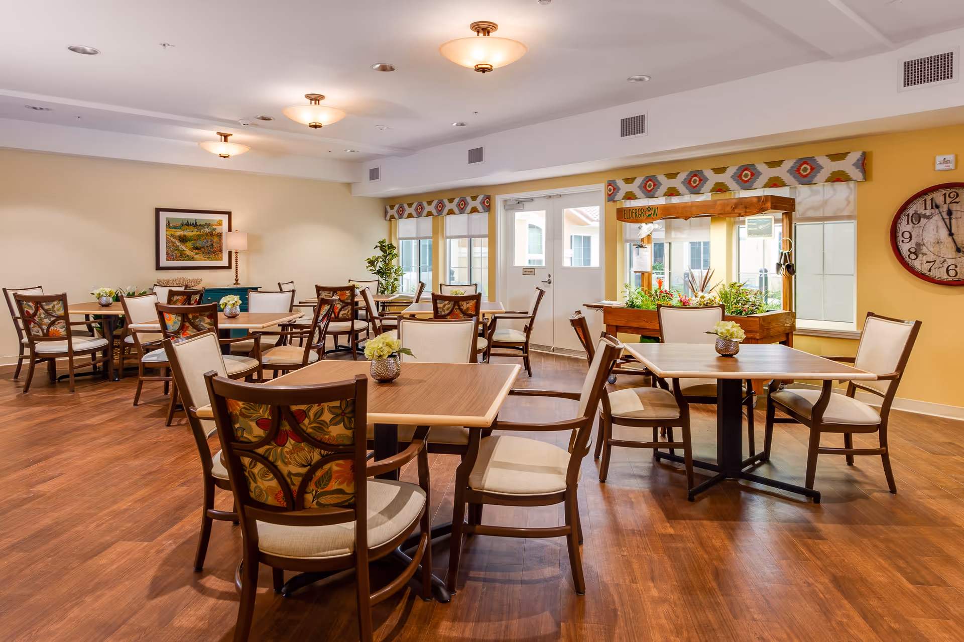 A bright and spacious dining room with multiple wooden tables and chairs arranged neatly. The chairs have cushioned seats and backs with a floral pattern. The room features wooden flooring, a large wall clock, a framed painting, potted plants, and a window with a decorative valance. There is also a small indoor garden or planter box near the windows and a door leading outside.