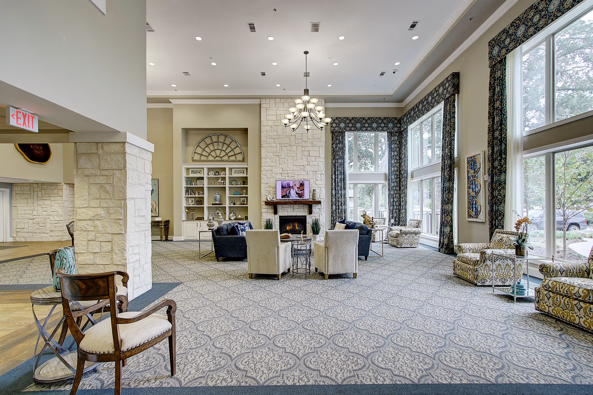 Spacious and well-lit living room area in a senior living facility with large windows, patterned carpet, multiple armchairs and sofas arranged around a stone fireplace with a TV mounted above it. The room features high ceilings with recessed lighting and a chandelier, decorative curtains, and built-in shelves with various decorative items.