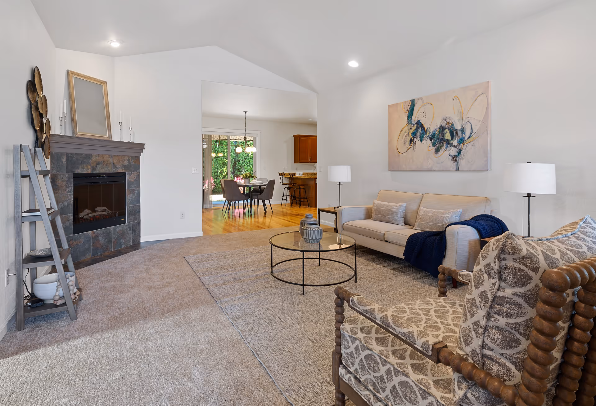 A cozy living room featuring a beige sofa with cushions and a navy throw, a patterned armchair, a round glass coffee table, two floor lamps, and a fireplace with a stone surround. The room has a neutral color palette with a large abstract painting on the wall and an open view into a dining area with a wooden table and chairs.