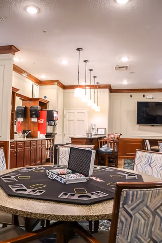 Interior room with a round table set up for poker with poker chips and card holders. The room has a kitchen area with a soda dispenser and cabinets, pendant lights hanging from the ceiling, and a wall-mounted TV. Several upholstered chairs surround the table and other seating areas are visible.