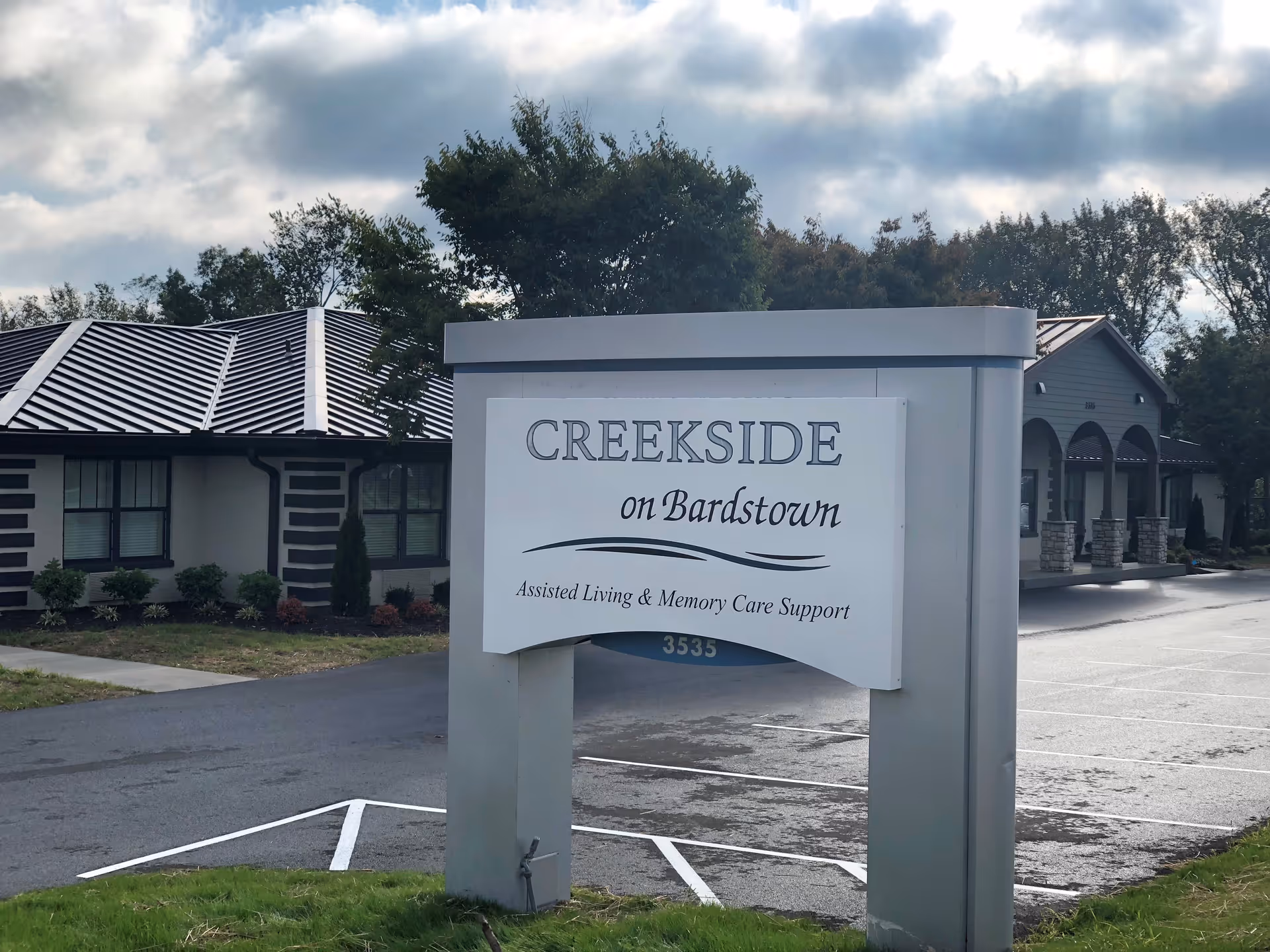 Outdoor view of the entrance sign for CreekSide on Bardstown, an assisted living and memory care support facility, with buildings and trees in the background under a cloudy sky.