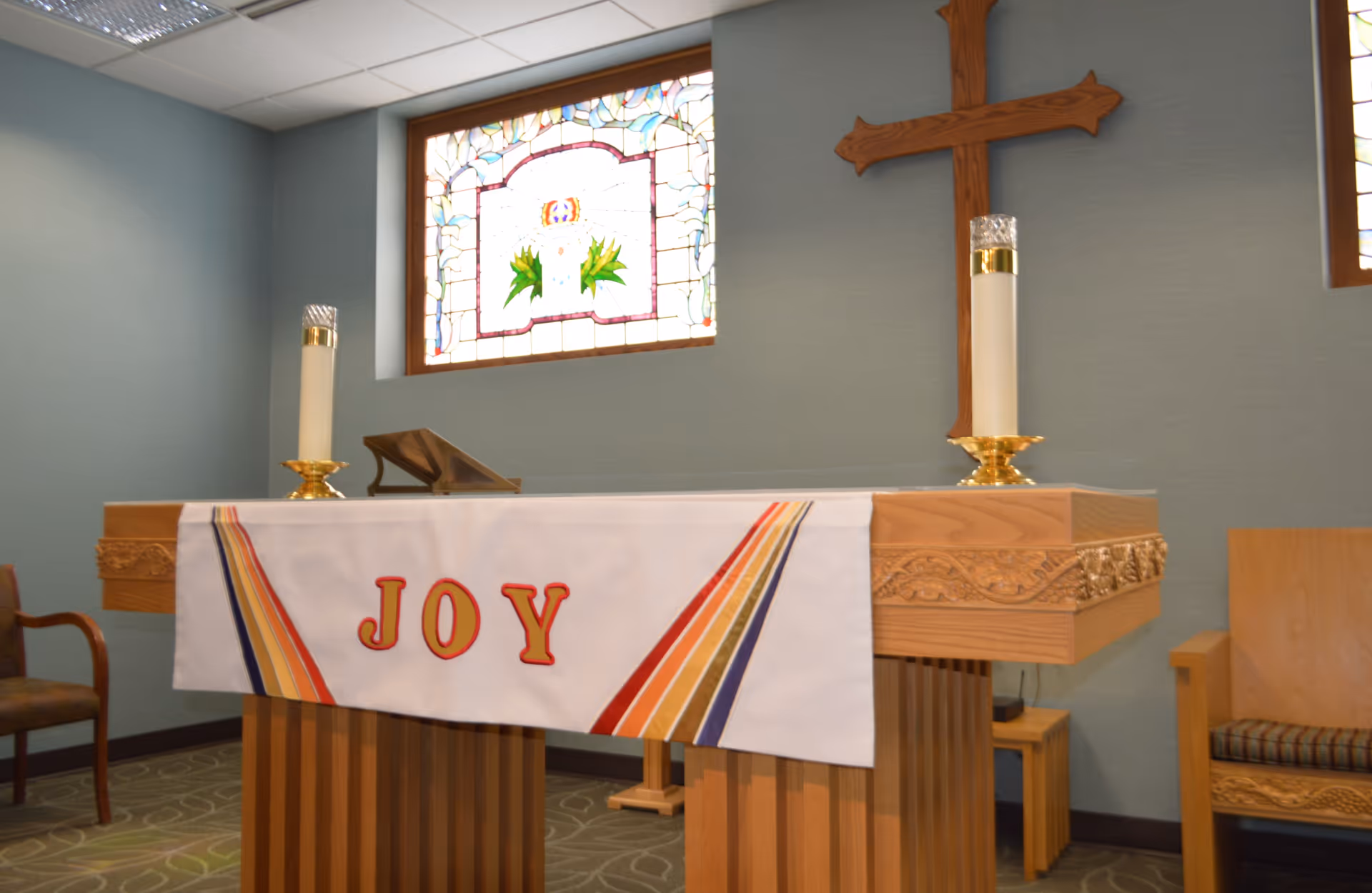Interior view of a small chapel or prayer room with a wooden altar draped with a white cloth that has the word 'JOY' in red letters and colorful stripes. Two tall candles in gold holders are placed on the altar. Behind the altar is a wooden cross mounted on a blue wall, and a stained glass window with floral designs is visible above the altar. Wooden chairs are positioned on either side of the altar.