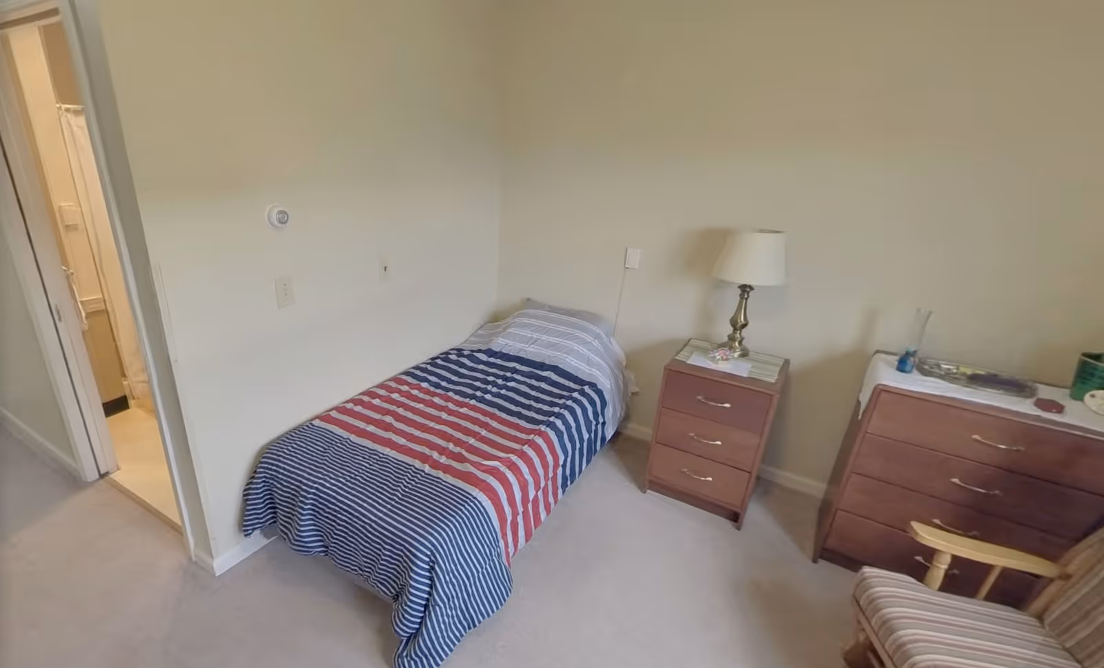 A small bedroom with a single bed covered in a striped red, white, and blue blanket. Next to the bed is a wooden nightstand with a lamp and some items on it. There is a wooden dresser with various objects on top and a wooden chair with striped upholstery. A doorway leads to a bathroom with a shower visible.