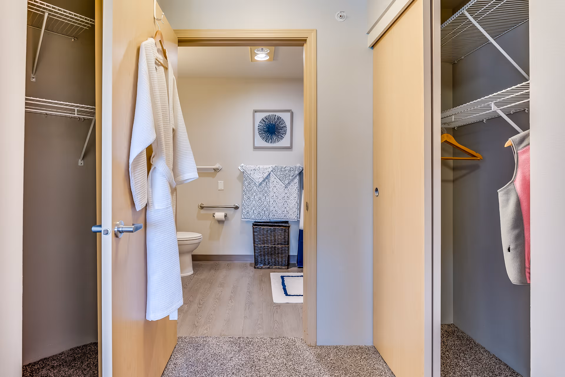 View through a hallway closet into a bathroom with a toilet, towel rack, laundry basket, and a robe hanging on the door.