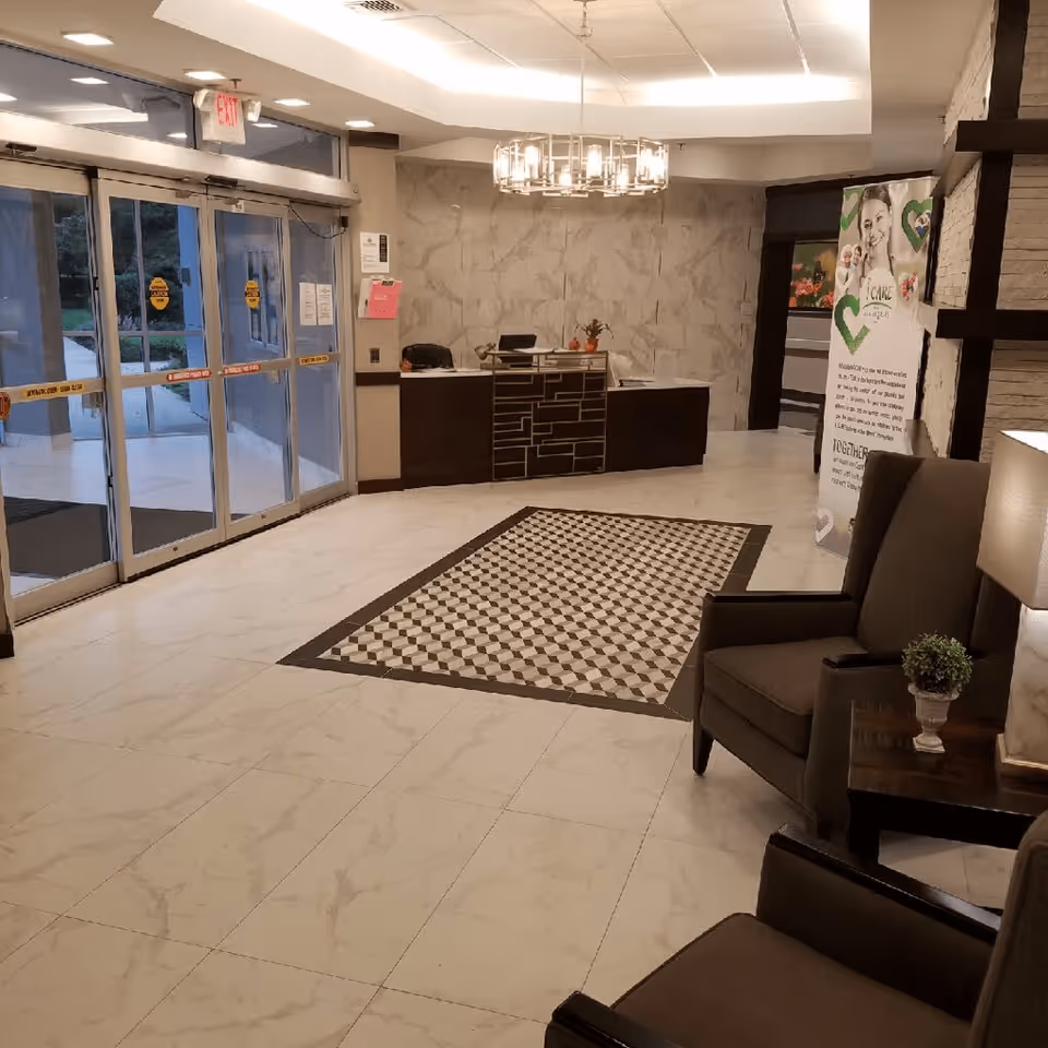 Interior view of a facility lobby with a reception desk, two armchairs, a side table with a lamp and plant, a patterned floor rug, and glass automatic sliding doors leading outside.