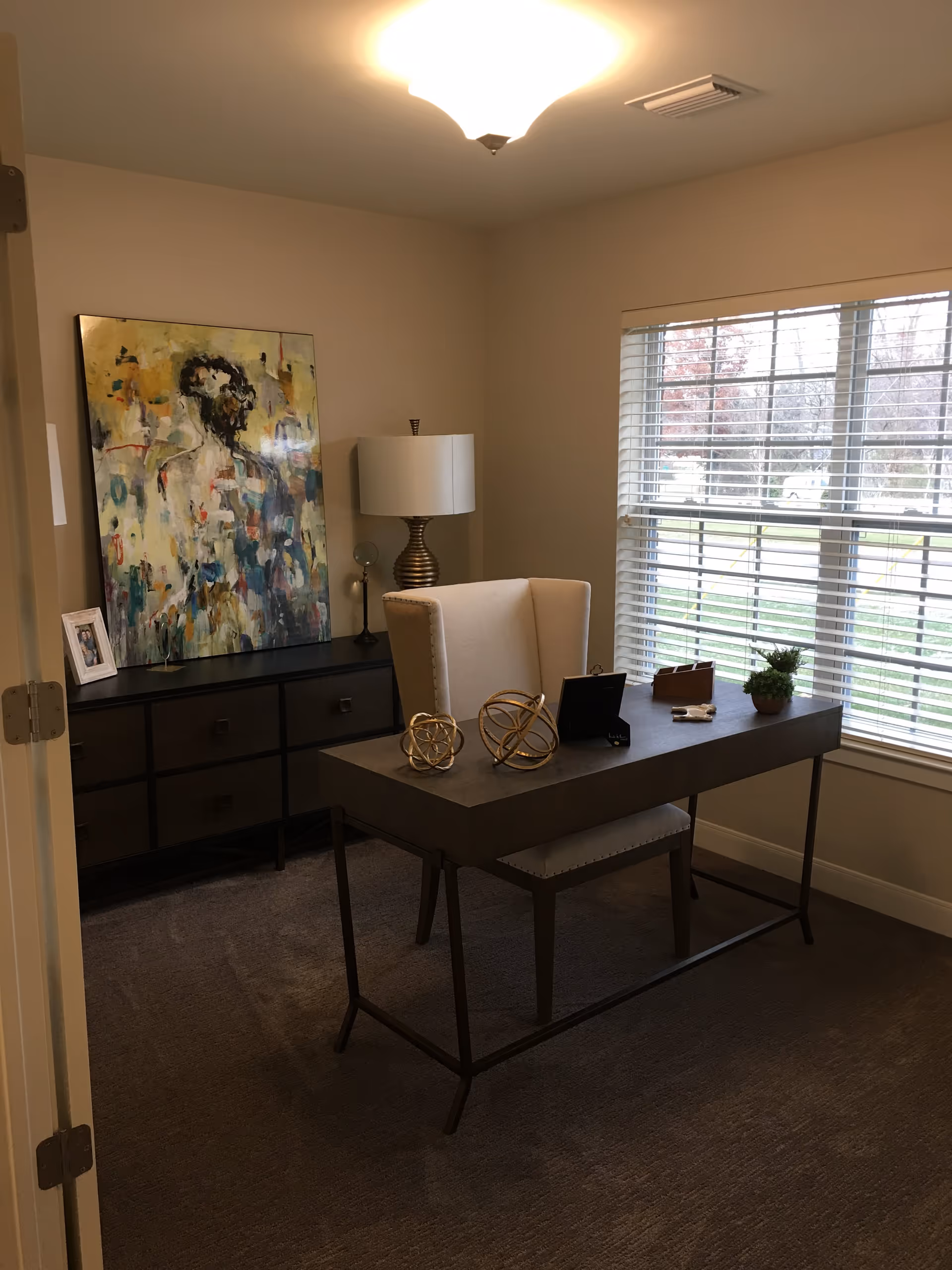 A small office or study room with a modern desk and a beige upholstered chair. On the desk are decorative items including two gold spherical sculptures, a small plant, and a few other objects. Behind the desk is a dark wooden dresser with multiple drawers, a table lamp with a white shade, and an abstract painting leaning against the wall. A large window with white blinds lets in natural light, showing a view of trees and grass outside.