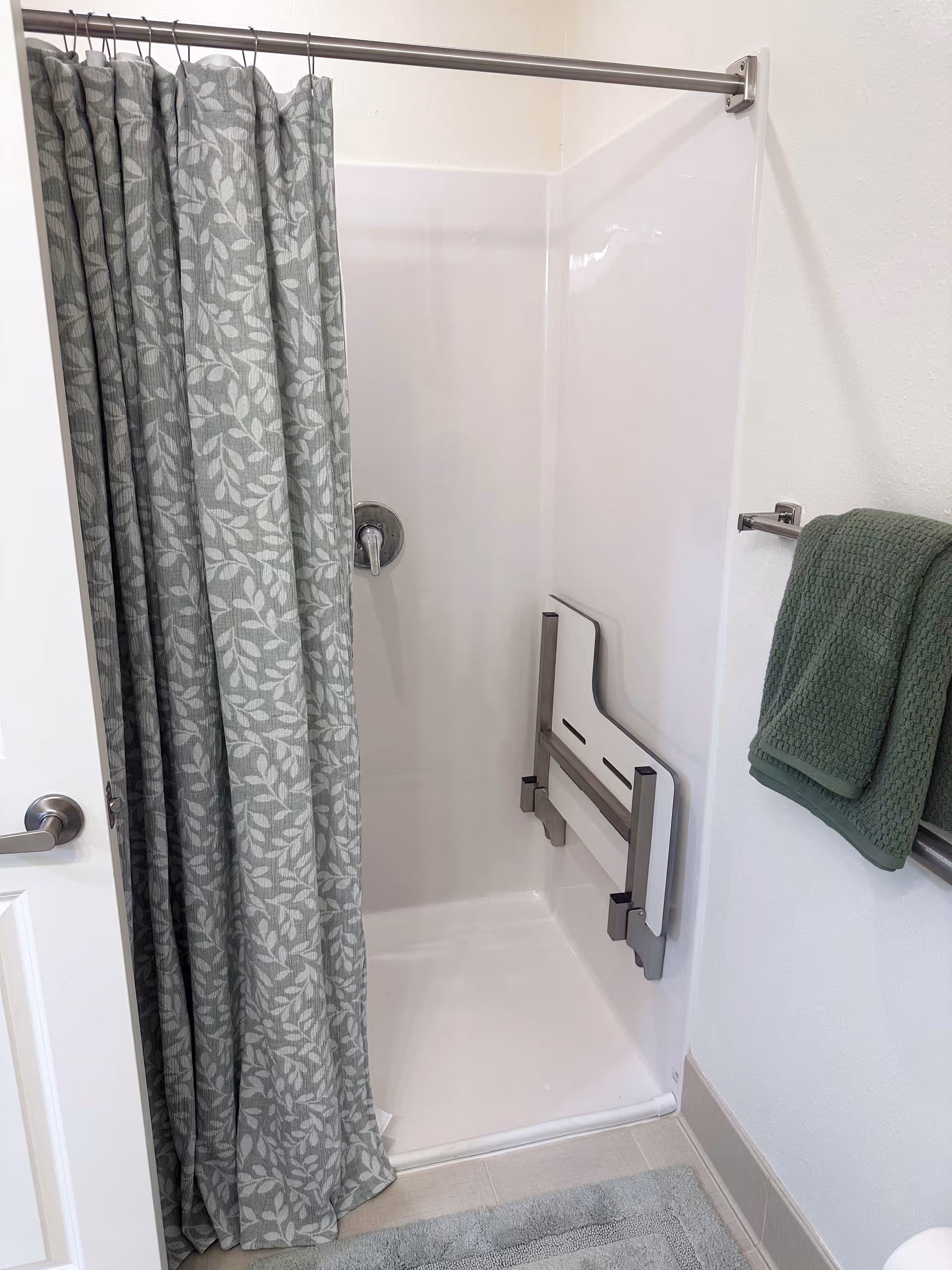 A small shower area with a gray and white leaf-patterned shower curtain partially drawn. Inside the shower, there is a foldable white shower seat mounted on the wall. A silver shower handle is visible on the left wall. Outside the shower, a green towel hangs on a silver towel rack attached to the white wall. The floor has a light-colored bath mat.