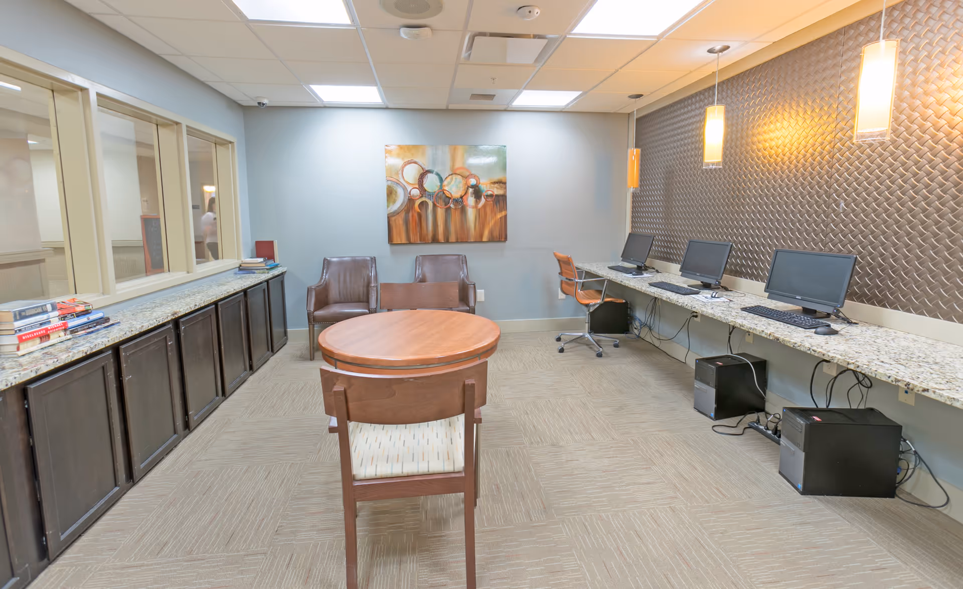 Interior common room with a round table and chairs, a row of desktop computers along a countertop, and seating against the far wall.