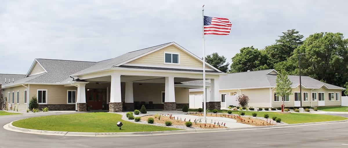 Exterior view of a single-story senior living facility building with beige siding and stone accents. The building has a covered entrance supported by white columns. In front of the entrance is a flagpole with an American flag. The surrounding area includes a circular driveway, landscaped grass, small bushes, and trees in the background under a cloudy sky.