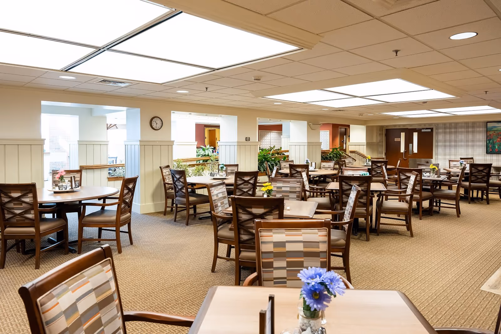 A spacious dining room with multiple wooden tables and chairs arranged neatly. Each table has a small vase with colorful flowers. The room has a beige carpet, white paneled walls, and a ceiling with large fluorescent lights. There are plants and a clock on the wall, with doors and windows visible in the background.