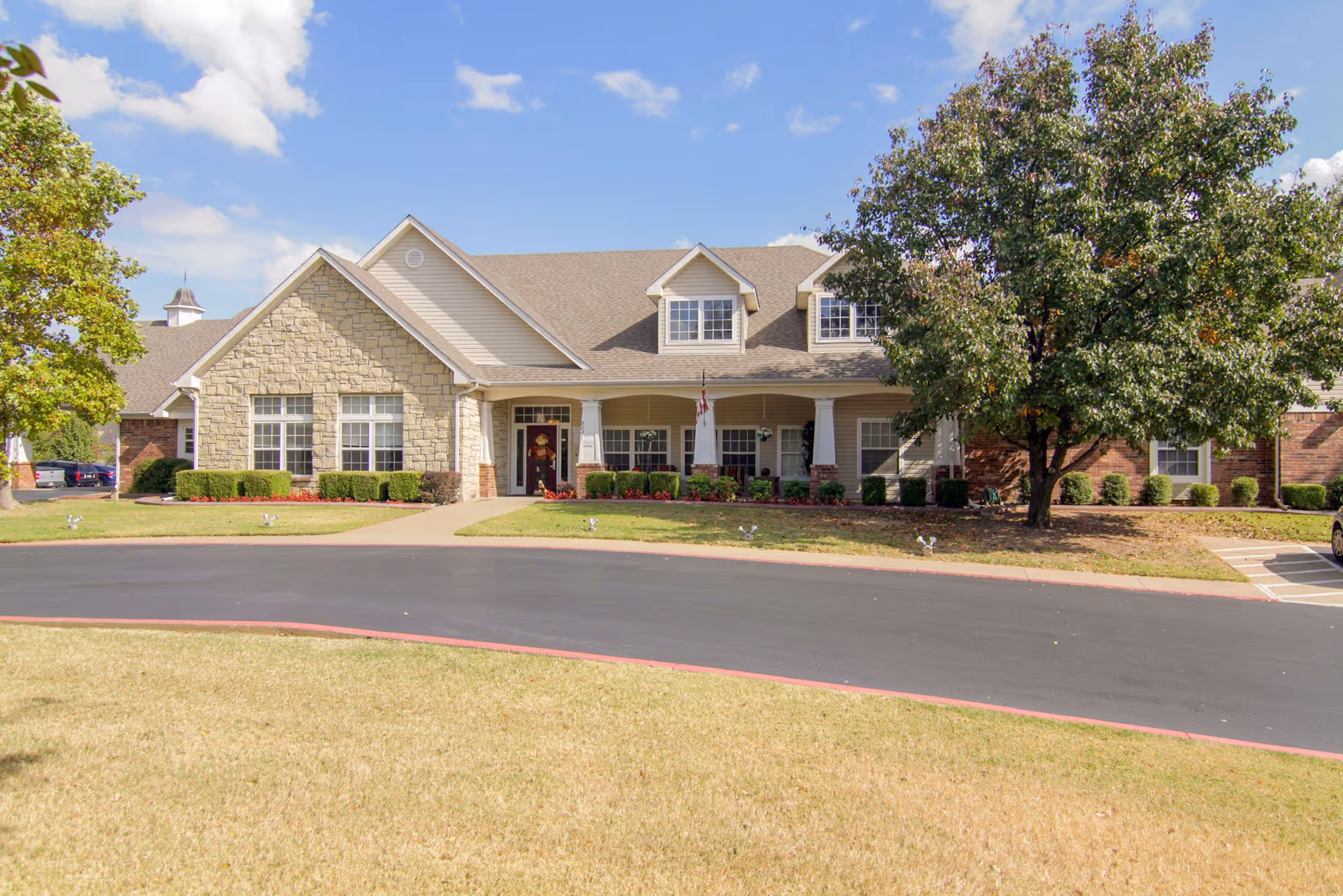 Front exterior view of Oxford Springs Tulsa Assisted Living facility showing a single-story building with a stone and brick facade, multiple windows, a covered entrance with white columns, surrounded by green bushes and trees under a partly cloudy blue sky.