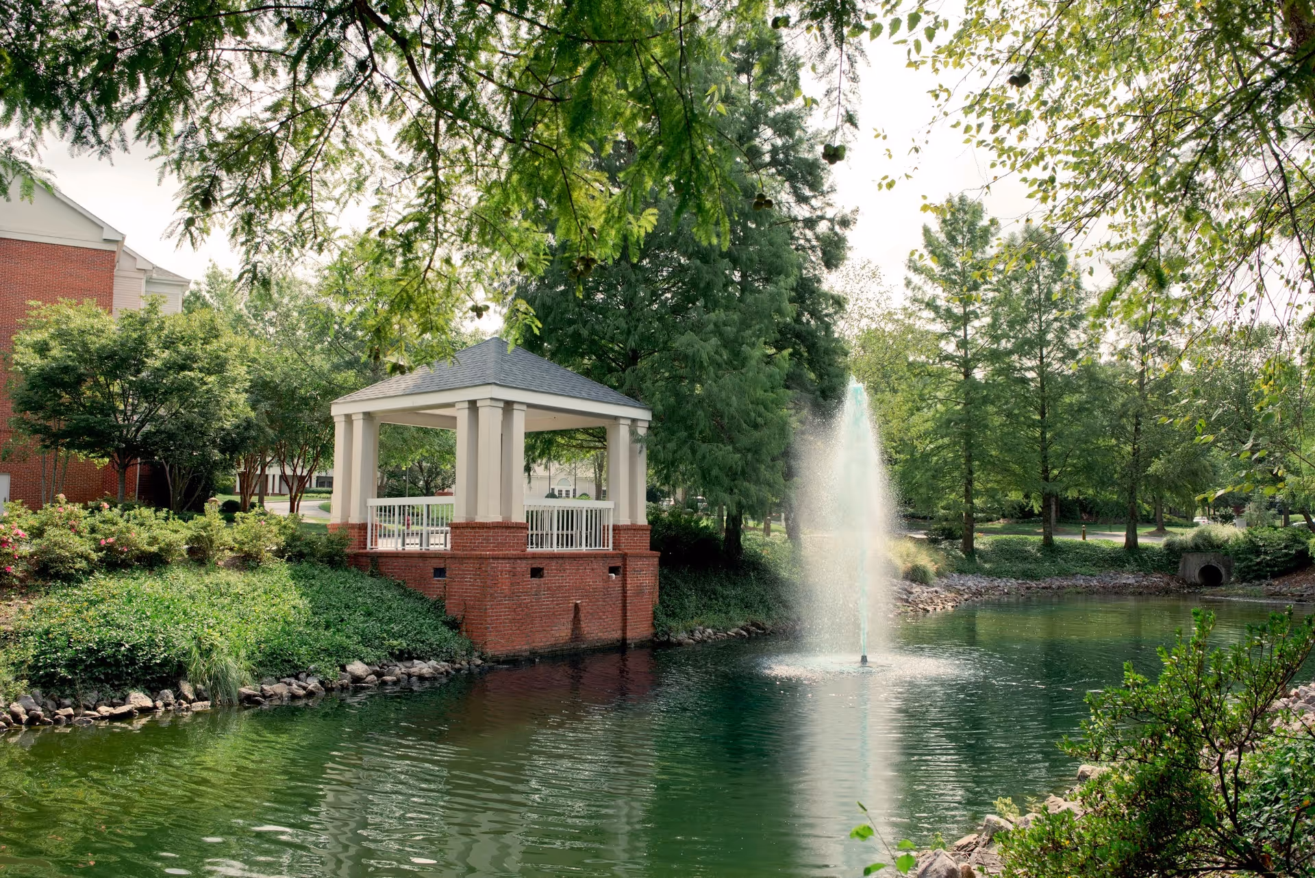 A serene outdoor scene at Royal Oaks featuring a small pond with a water fountain in the center, surrounded by lush green trees and shrubs. There is a white gazebo with a brick base on the edge of the pond, and a red brick building partially visible in the background.