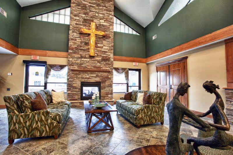 A cozy living room area with two patterned sofas facing each other across a wooden coffee table. The room features a tall stone fireplace with a large wooden cross mounted above it. There are windows on either side of the fireplace with curtains, and the walls are painted green and beige with wooden trim. In the foreground, there is a sculpture of two abstract human figures holding hands.