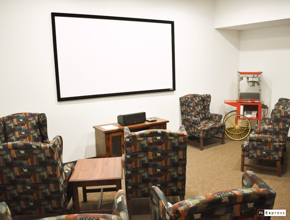 A small theater or media room with patterned armchairs arranged in rows facing a large blank projection screen on the wall. There is a wooden cabinet with a speaker and remote control in front of the screen, and a red popcorn machine on a stand with a yellow wheel next to it in the corner.