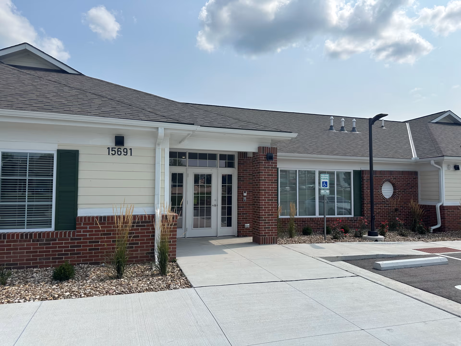 Front exterior view of a single-story building with a combination of brick and light-colored siding. The entrance features double glass doors with sidelights and a small covered porch. There are windows with white blinds and green shutters, a handicapped parking sign, and a streetlamp near the entrance. The building number 15691 is displayed on the wall near the door. The sky is partly cloudy.