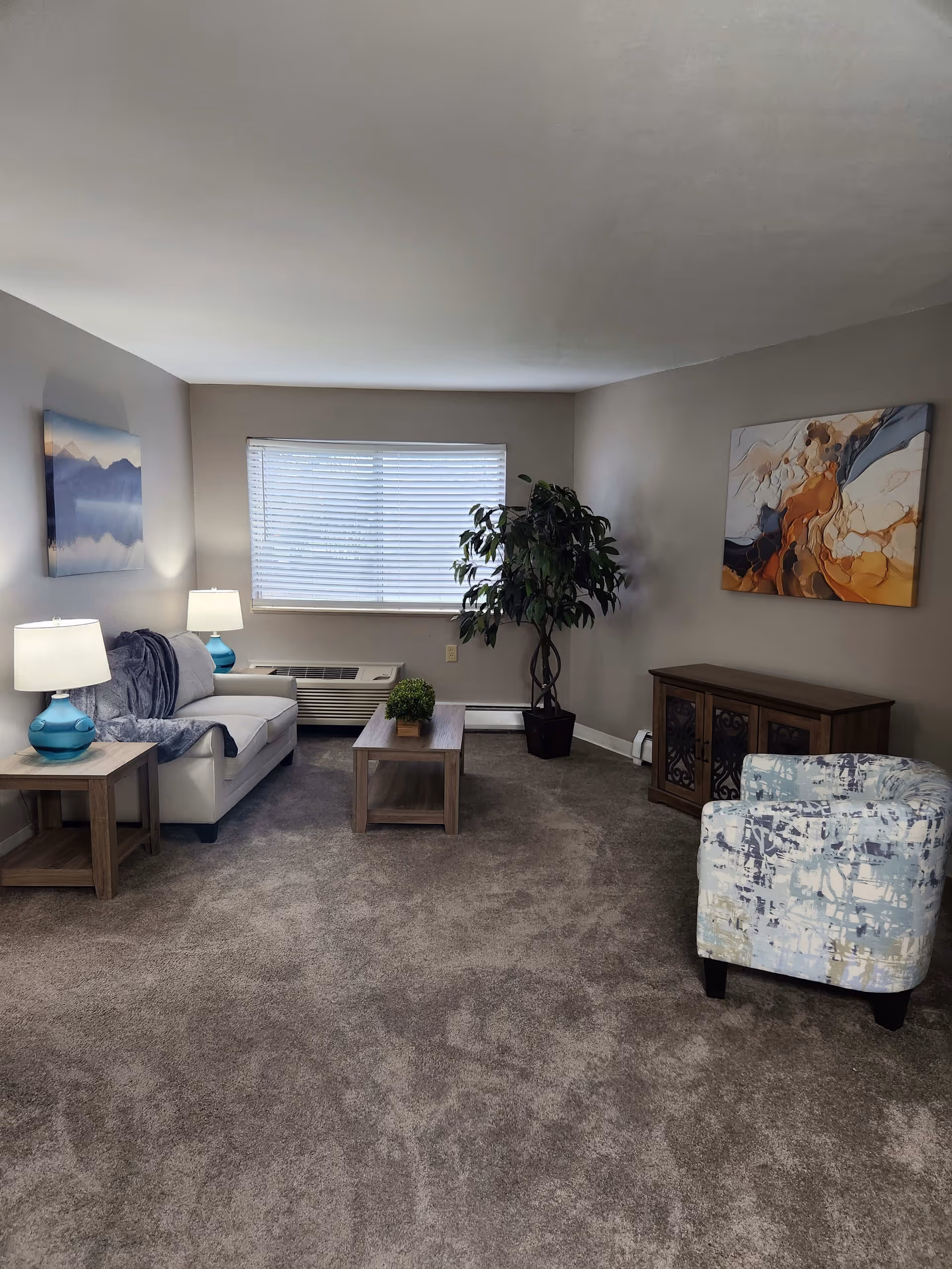 A living room with beige walls and carpeted floor featuring a white sofa with a blue throw blanket and two blue lamps on wooden side tables. There is a wooden coffee table with a small green plant, a large potted plant near the window with blinds, a patterned armchair, a wooden cabinet, and two abstract paintings on the walls.