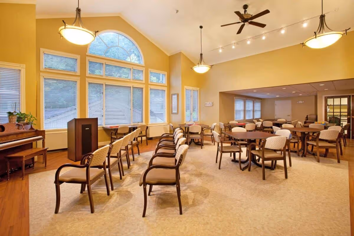 Spacious senior living common room with rows of chairs, round dining tables, a piano, large arched windows, and warm overhead lighting.